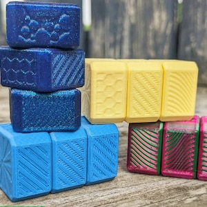 May include: A stack of colorful, patterned, cube-shaped fidget toys. The top layer is yellow, the middle layer is blue, and the bottom layer is blue with a white pattern.