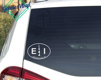 May include: White oval sticker on a car window featuring the letters "E I" and a lighthouse design. The text "EMERALD ISLE, NC" is below the letters. The sticker is a travel souvenir.