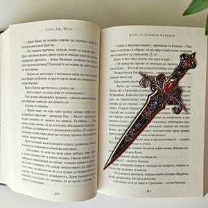May include: A close-up of a book with a bookmark shaped like a sword. The bookmark is made of metal and has a red gem on the handle. The book is open to a page with text in a foreign language. The text on the page reads "Dvor ot Sreburni Plamaci" and "Spri s dvusmislicitie - prekusna go Kasidi. Tam din e ostanal v obraza si na zvar i nai-setne poluchava zaslujenoto si nakazanie. E. i"