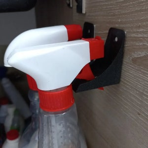 May include: A black spray bottle holder mounted on a wooden surface. The holder is designed to hold two spray bottles. The spray bottles have white and red tops. The holder is made of black plastic.