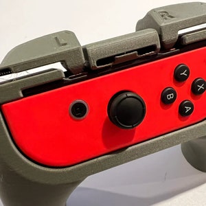 May include: A gray 3D printed case for a Nintendo Switch controller. The case has a red controller inside with black buttons. The case has a textured surface and a raised lip around the edge.