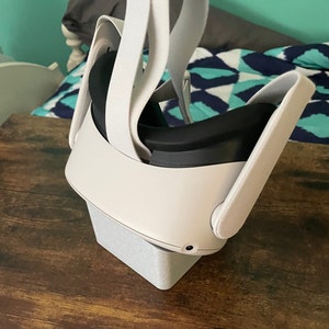 May include: A white virtual reality headset with black interior padding and adjustable white straps. The headset is resting on a light gray, rectangular stand, set against a wooden surface.