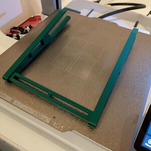 May include: A green plastic frame sits on a textured, light brown surface. The frame has a rectangular shape with a grid pattern inside.