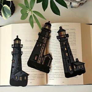 May include: Three black and gold lighthouse bookmarks with intricate details. The bookmarks are placed on an open book with Russian text.