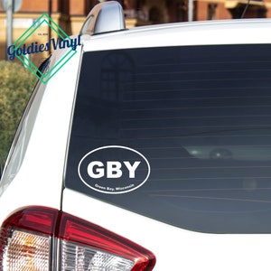 May include: White oval sticker with the text "GBY" and the words "Green Bay, Wisconsin" underneath. The sticker is on the back window of a white car.