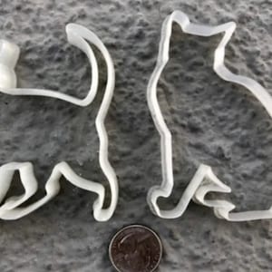 May include: Two white plastic cat-shaped cookie cutters. The cutters are in the shape of a cat sitting with its tail curled around its body.