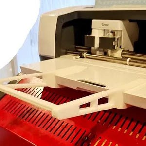 May include: A white plastic accessory for a Cricut cutting machine. The accessory is a shelf that attaches to the top of the machine. The shelf is designed to hold materials while cutting.