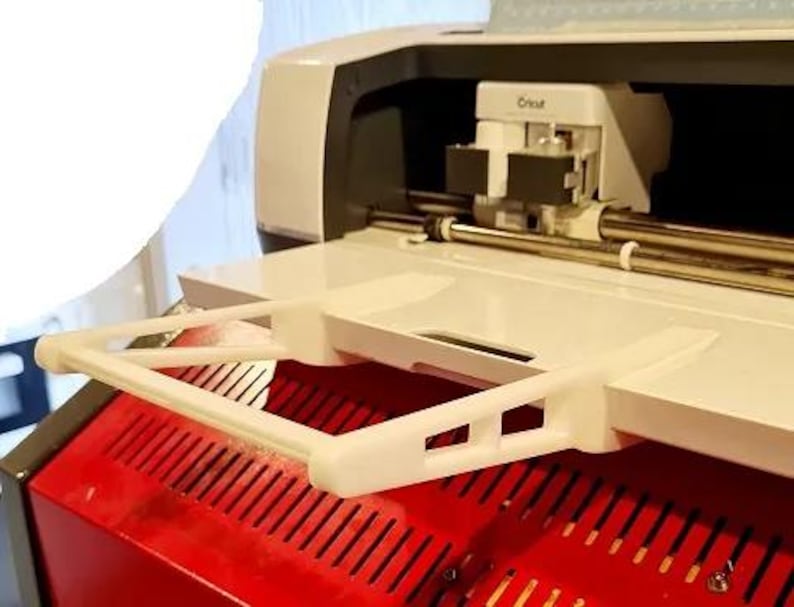 May include: A white plastic accessory for a Cricut cutting machine. The accessory is a shelf that attaches to the top of the machine. The shelf is designed to hold materials while cutting.