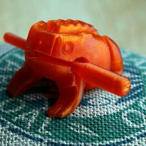 May include: A vibrant orange, 3D-printed object with a unique design. It features a central, textured body with a cylindrical rod passing through it. The object rests on a patterned fabric, showcasing its intricate details and form.