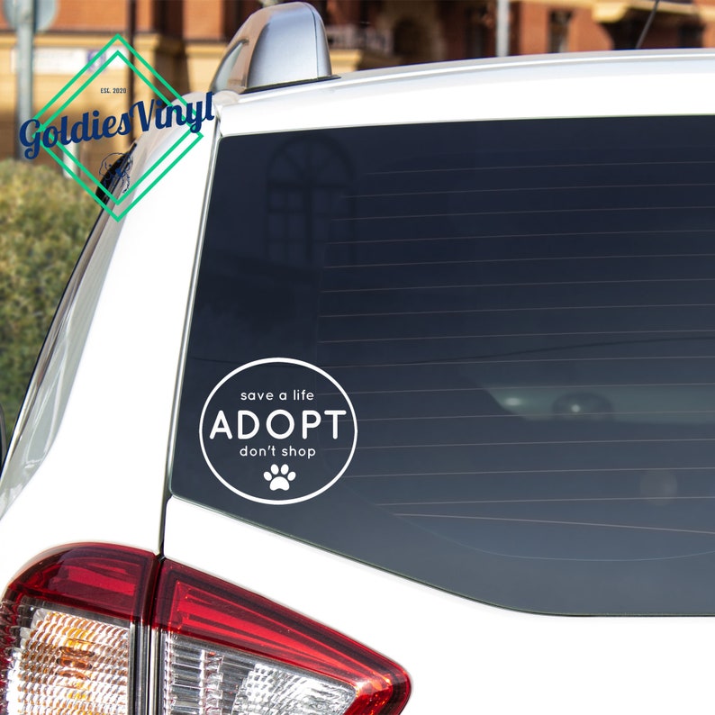 Op de afbeelding: Witte auto-raamsticker met een cirkelvormig ontwerp met de tekst "red een leven ADOPTEER koop niet" met een pootprint-icoon.