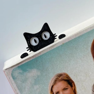 May include: A black cat bookmark with white eyes peeking over the top of a book. The book has a photo of two women on the cover.