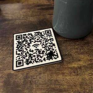May include: A white square coaster with a black and white QR code featuring a wifi symbol in the center.