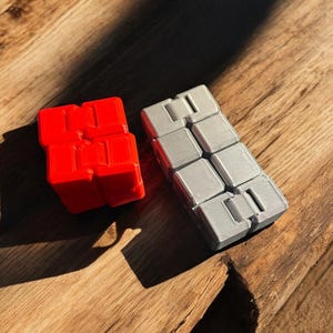 May include: Two fidget toys, one red and one silver, made of plastic. The toys are made up of small squares that can be flipped and rotated.