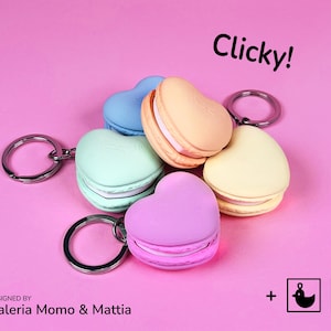 May include: Heart-shaped keychains in pastel colors, including blue, green, peach, and yellow, with silver key rings. The word "Clicky!" is written in black. Designed by Valeria Momo & Mattia.