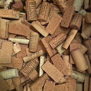 May include: A close-up image of a pile of used wine corks. The corks are various sizes and shapes, with some having the names of wineries printed on them. The corks are a mix of brown, tan, and beige colors.