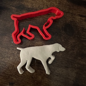 May include: A red plastic cookie cutter shaped like a dog, with a white clay cutout of a dog inside.
