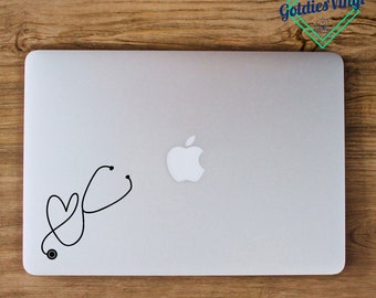 May include: A white laptop computer with a black stethoscope and heart design sticker on the lid. The Apple logo is visible on the lid. The laptop is on a wooden surface. *laptop not included*