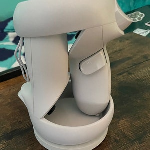 May include: A white VR headset and controllers are docked on a white charging station. The controllers are positioned on either side of the headset, with the charging station base resting on a wooden surface. The setup is designed for virtual reality gaming.