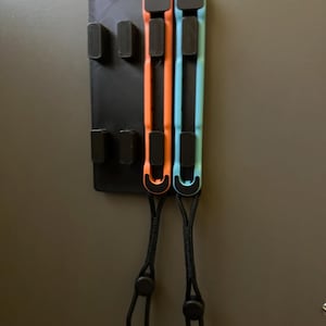 May include: A black wall-mounted organizer with slots and two colorful straps. The straps are orange and blue, with black cords and adjustable toggles. The organizer is mounted on a gray wall.
