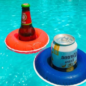 May include: Two inflatable drink holders, one orange and one blue, float in a pool. An amber beer bottle sits in the orange holder, and a silver can of beer rests in the blue holder. The water is a bright turquoise color.