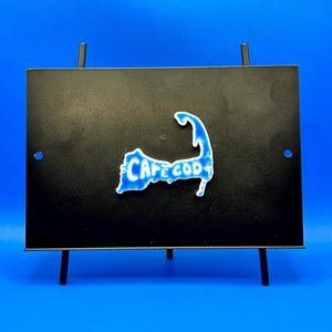 May include: A black metal display stand with a blue and white Cape Cod map design. The map is affixed to a black rectangular surface, supported by four thin legs. The background is a solid blue.