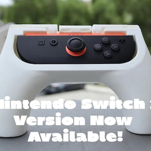 May include: White Nintendo Switch 2 controller grip with a black controller and orange accents. The text "Nintendo Switch 2 Version Now Available!" is displayed on the grip.