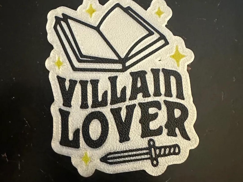 May include: A white sticker with the words "Villain Lover" in black, stylized font. The sticker features an open book, a dagger, and yellow star accents. The sticker has a textured surface.