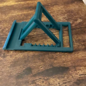 May include: A dark green plastic phone stand with a simple design. The stand is folded up and ready to be used.