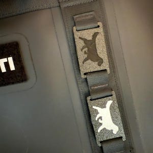 May include: Close-up of a black cooler bag with a black strap and two rectangular metal buckles. Each buckle features a silhouette of a goat. The bag has a black patch with white text.