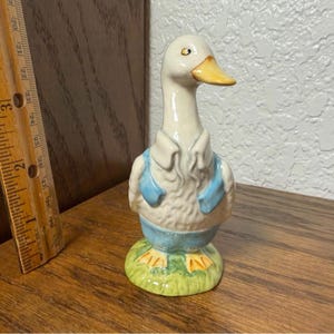 May include: A ceramic duck figurine with a white body, yellow beak, and blue overalls. The duck stands on a green base with yellow feet. The figurine is approximately 5 inches tall and is a decorative item.