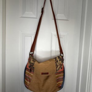 May include: A crossbody bag featuring a tan textured center panel, side panels with a multi-colored woven pattern, and a dark blue denim base. The bag has a long brown adjustable strap and a small rectangular red label on the front flap.