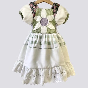 May include: A handmade white and green patchwork dress with a floral design. The dress features a large flower applique on the bodice, puff sleeves, and a gingham skirt. The hemline is finished with a scalloped eyelet trim.
