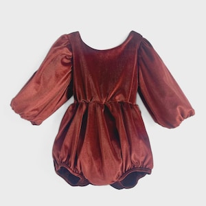 May include: A rust-colored velvet romper with long, puffed sleeves and a rounded neckline. The romper features a gathered waist and a bubble-style bottom, creating a vintage-inspired look. The fabric has a soft sheen, suggesting a luxurious feel.