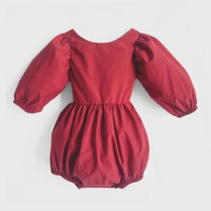 May include: A red baby romper with long, puffy sleeves and a gathered waist. The romper has a round neckline and a bubble-style bottom. The fabric appears to be a soft, solid-colored material.