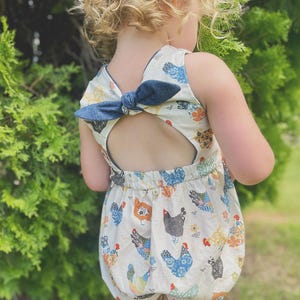 May include: A sleeveless romper with a back cutout and a denim bow. The romper features a cream background with colorful chicken illustrations. The design includes various chicken breeds in blue, orange, and black.