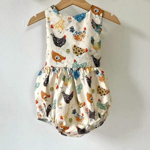 May include: A cream-colored baby romper with a playful chicken print in various colors. The sleeveless romper features a round neckline and a gathered, bubble-style bottom. It hangs on a wooden hanger against a plain white background.