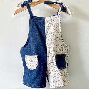 May include: A split-design romper with denim and floral fabric. The denim side is a dark blue, while the other side features a white background with a small floral pattern. The straps are tied with floral fabric bows. Two pockets are visible.