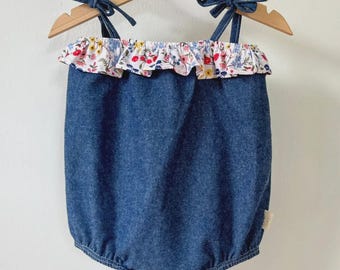 Puede incluir: Un pelele de bebé de mezclilla azul con un ribete de volantes florales. El pelele tiene tirantes anudados y está colgado de una percha de madera. El ribete floral presenta flores rojas, rosas y amarillas sobre un fondo blanco.