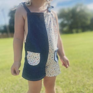 Op de afbeelding: Een tweekleurige romper voor peuters. De ene kant is donkerblauwe denim, de andere is wit met een bloemenpatroon. De romper heeft een zak aan de denimkant en strikbandjes. Het kind staat op groen gras.