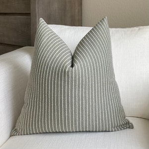 May include: A gray decorative pillow with vertical white stripes. The pillow is placed on a white sofa. The pillow has a textured appearance and a soft, inviting look. The pillow is a square shape.