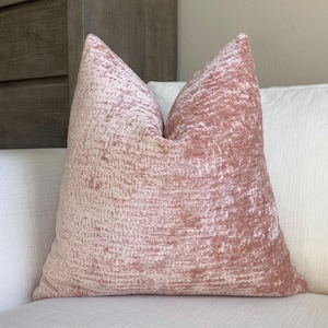 May include: A blush pink decorative pillow with a textured, woven appearance. The pillow is square and has a soft, plush look. The pillow is resting on a white surface.