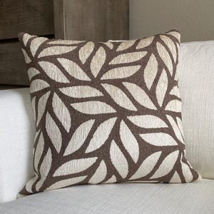 May include: Square decorative pillow with a brown and beige leaf pattern. The pillow is placed on a white sofa. The pillow's design features a repeating pattern of stylized leaves in a neutral color palette.