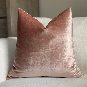 May include: A blush pink velvet pillow with a soft, plush texture. The pillow is shown on a white couch.