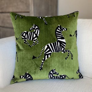 May include: Square green decorative pillow with a repeating pattern of black and white zebras. The pillow has a soft, velvety texture and is placed on a white sofa. The zebras are depicted in various poses, adding a dynamic element to the design.