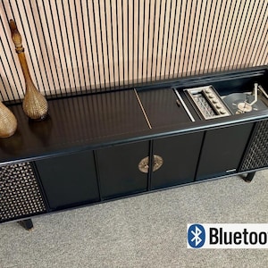 May include: A black wooden media console featuring a built-in record player under a sliding top panel, two side speaker grilles with a woven texture, central cabinet doors with circular brass hardware, and two tall amber glass decorative bottles.