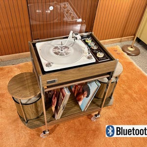 May include: A Zenith record player console with a wood grain finish, featuring a white turntable, a transparent hinged lid, and a lower shelf holding vinyl records. The unit is mounted on a rolling metal frame with two circular side stools.