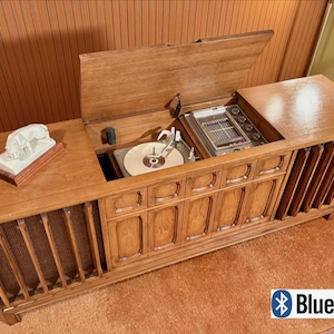 May include: A long wooden console stereo cabinet with a hinged top lid revealing a built-in record player and radio tuner. The unit features vertical slatted speaker grilles on the sides and a white animal figurine on the left surface area.