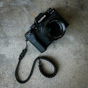 May include: A black Fujifilm X-T5 camera with a black lens and a black camera strap. The camera has a black strap with a black buckle.