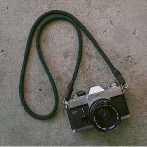 May include: A dark green camera strap with a silver Canon camera attached. The camera has a black lens with the text "Canon FD 50mm 1:1.8 SC" printed on it.