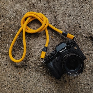 Könnte beinhalten: Eine schwarze Fujifilm X-T3 Kamera mit einem gelben Kameragurt aus Seil. Das Objektiv der Kamera ist nach vorne gerichtet. Der Gurt besteht aus einem dicken, gewebten Material. Die Kamera liegt auf einer strukturierten, grauen Oberfläche.